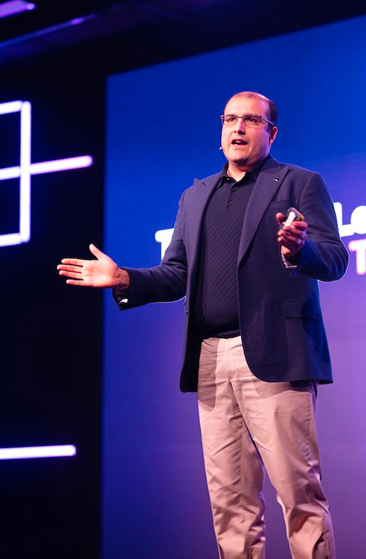 Vishal Marria speaks on stage with his hands outstretched, under bright stage lighting.