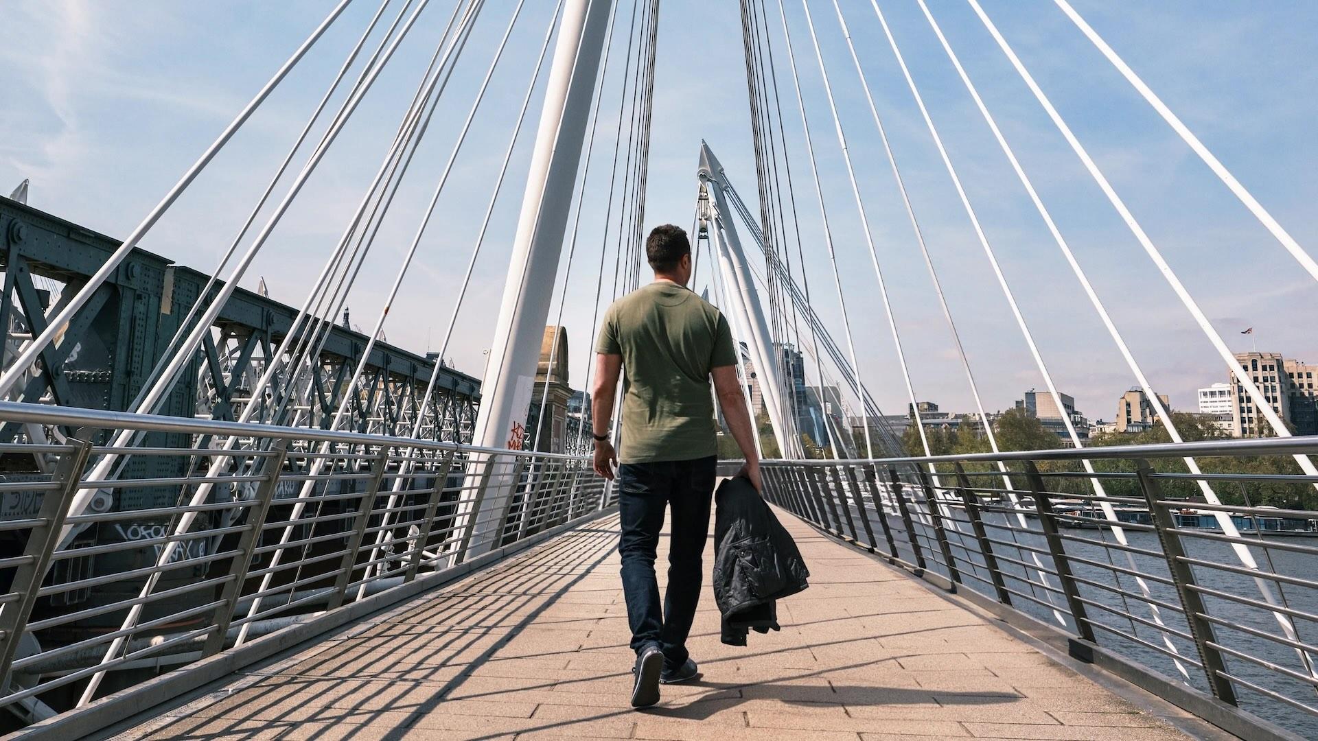 A man walks on a modern suspension bridge, holding a jacket, with city buildings visible in the background under a clear sky.