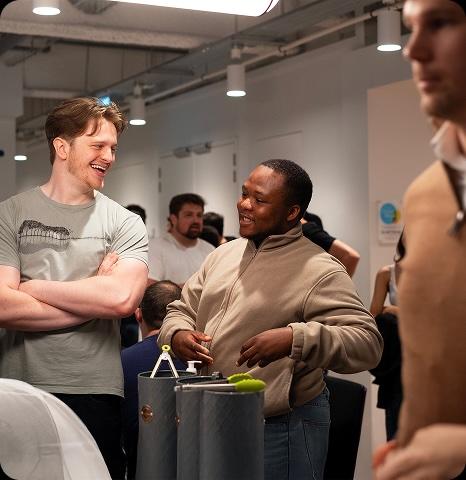 Two people are smiling and conversing in an office setting. They are surrounded by colleagues and office equipment.