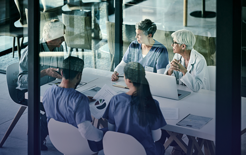 Healthcare professionals in a meeting room, discussing documents and using a laptop, viewed through a glass wall.