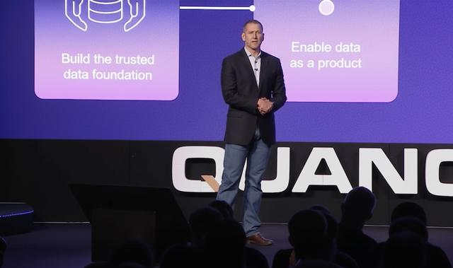 Man in a dark blazer talks on stage, standing in front of a screen displaying tech-related text and diagrams.