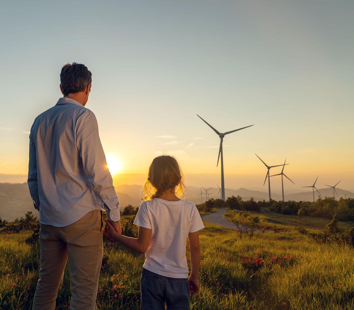 Man and child holding hands, facing wind turbines during a sunset on a grassy hill.