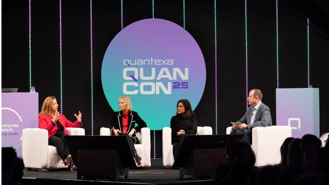 Four people sit on a stage panel discussing at Quantexa QUANCON25, with stage lights and a large logo in the background.